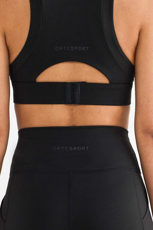 The individual is wearing a black ORTC Sport outfit, including the ORTC Eva Crop Top and leggings. The image showcases the back clasp and design of the Eva Crop Top, which provides firm support with its brushed fabric for maximum comfort.