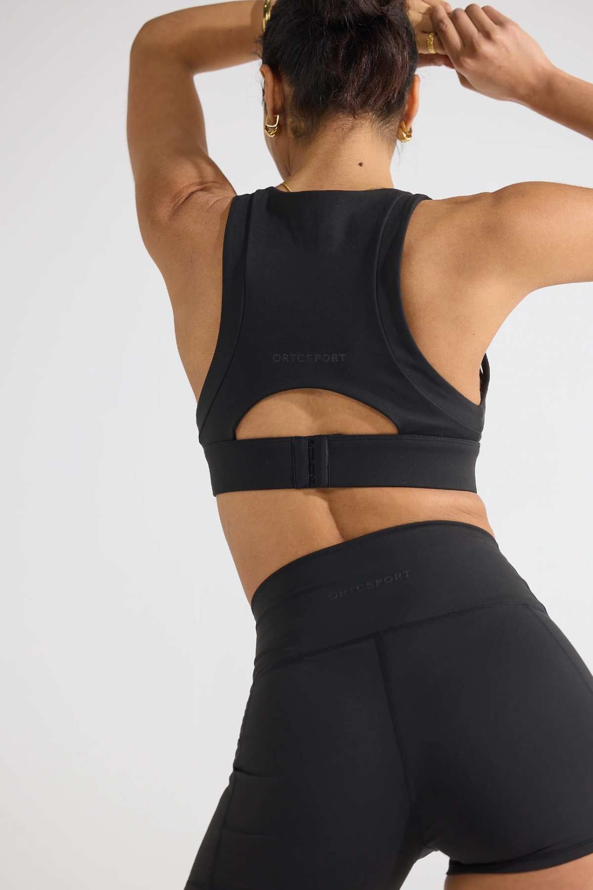 A person facing away, wearing a black ORTC Eva Crop Top and brushed fabric shorts, with their hair tied up. They are in a stretching pose with arms raised, offering firm support against a plain background.