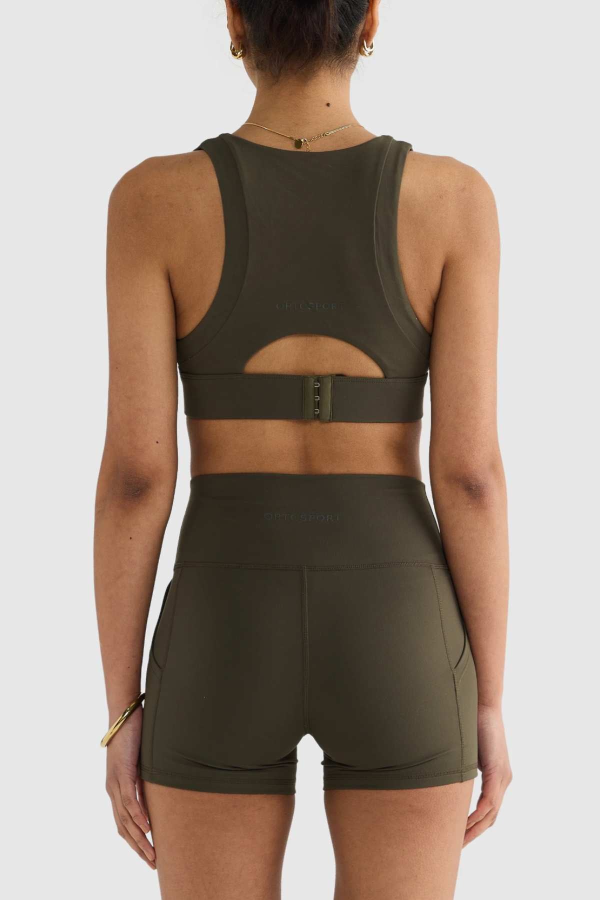 A person wearing the ORTC Eva Crop Top, a dark green athletic sports bra and shorts set crafted from recycled polyester for an eco-friendly touch, stands with their back to the camera. The back strap design provides firm support, making it ideal for any workout. The background is plain and light-colored.