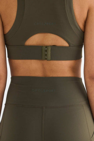 A person wearing a matching olive green ORTC Sport set, which includes the Eva Crop Top and leggings made from recycled polyester. The firmly supportive crop top features a hook clasp and a small cutout on the back, with the brand name visible on both pieces.