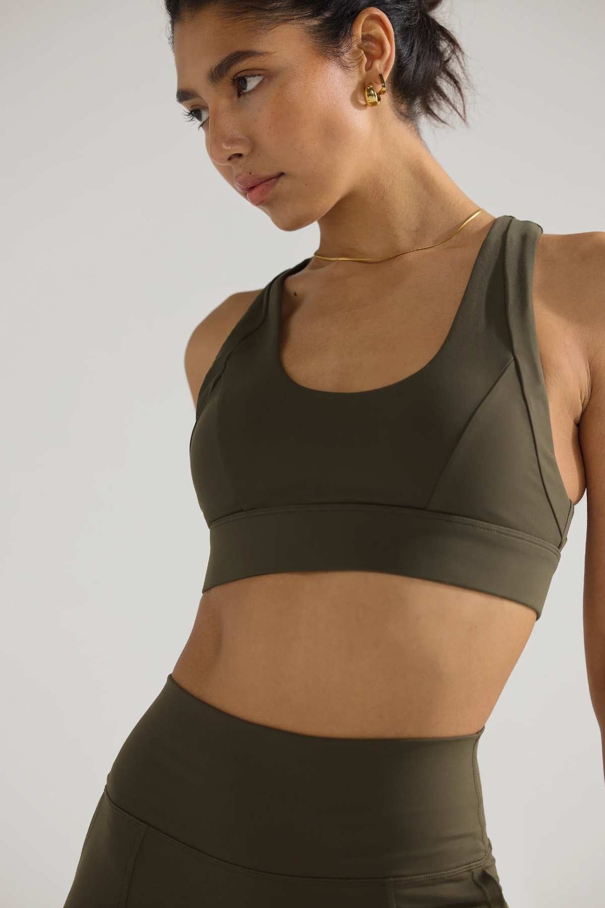 A woman wearing the ORTC Eva Crop Top in dark olive, paired with matching leggings made of recycled polyester, looks to the side. Her hair is tied back, and she accessorizes with gold hoop earrings and a necklace against a plain, light-colored wall.