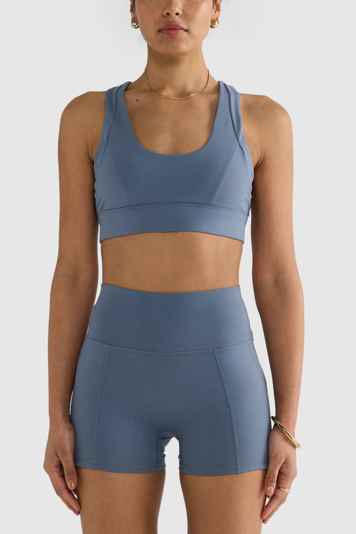 A person wearing a blue ORTC Eva Crop Top paired with matching high-waisted shorts stands against a plain white background. They have short hair and are accessorized with a gold necklace and bracelet, exuding confidence in their outfit's firm support and durable fabric.