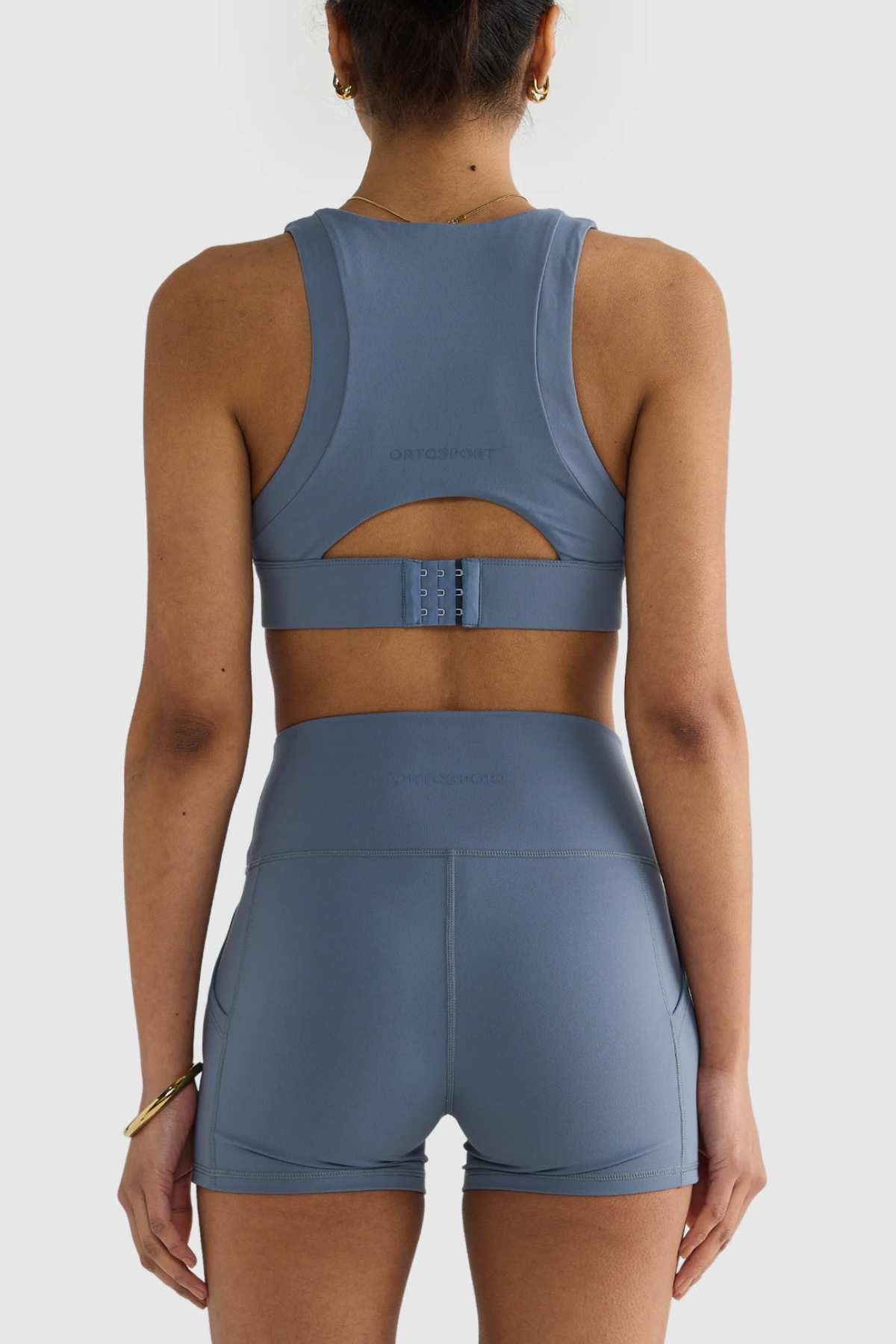 Rear view of a person wearing an ORTC Eva Crop Top, paired with matching high-waisted shorts, providing firm support and made from durable fabric. The sophisticated ensemble is enhanced by wrist jewelry, set against a plain, neutral backdrop.