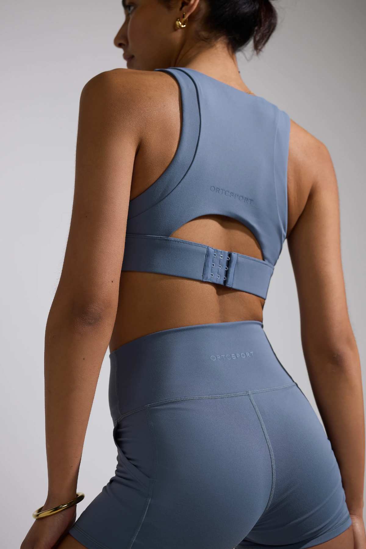 A person is wearing an ORTC Eva Crop Top, perfectly paired with high-waisted shorts in blue. Captured from the back, the outfit showcases its durable fabric and subtle branding. The individual, with dark hair tied up, completes the look with gold hoop earrings and a bracelet.