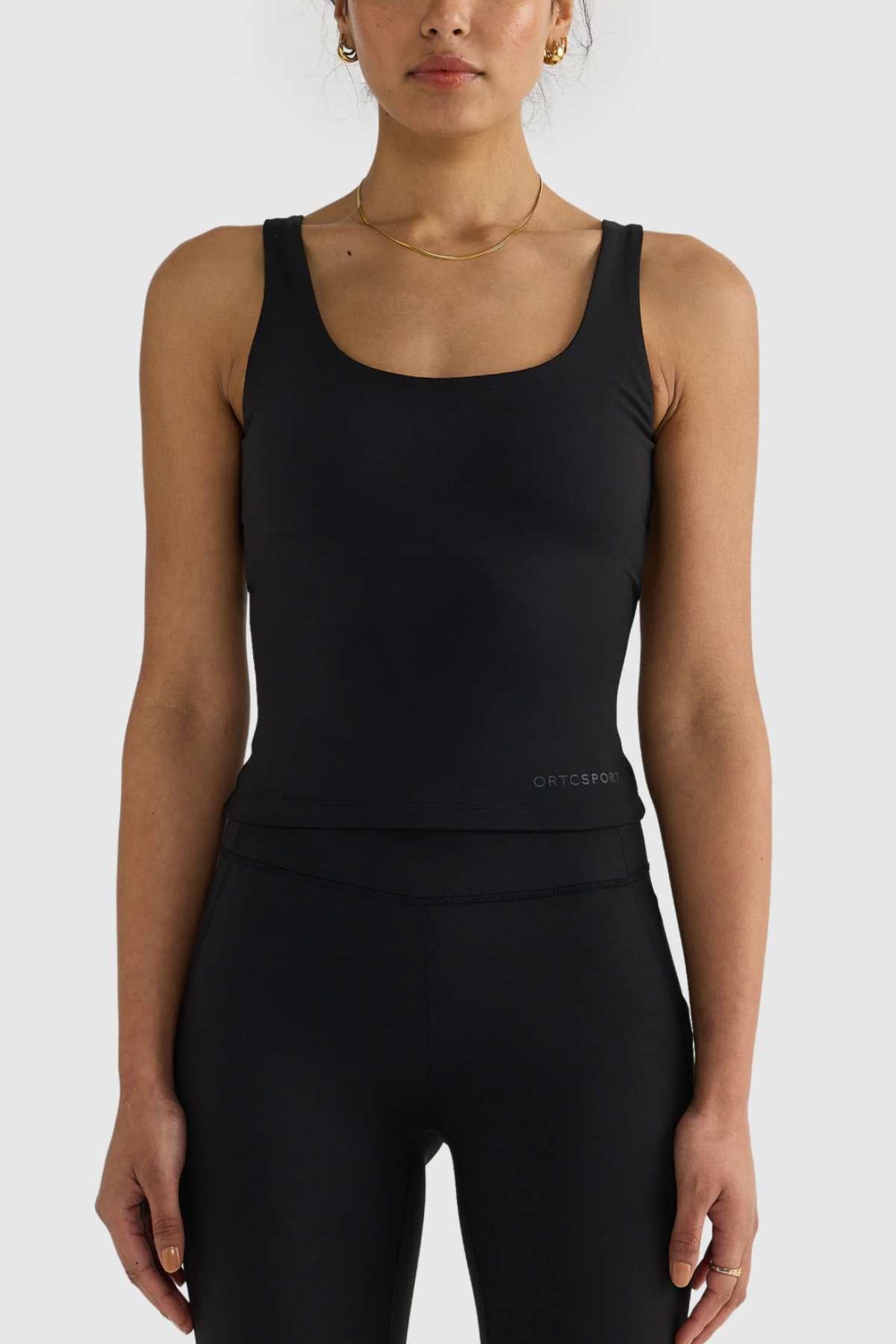 A person stands against a plain background wearing the ORTC Eva Twist Back Reversible Top in black along with matching black leggings. They accessorize with gold hoop earrings and a delicate necklace. The ensemble is form-fitting and sporty, ideal for versatile styling.