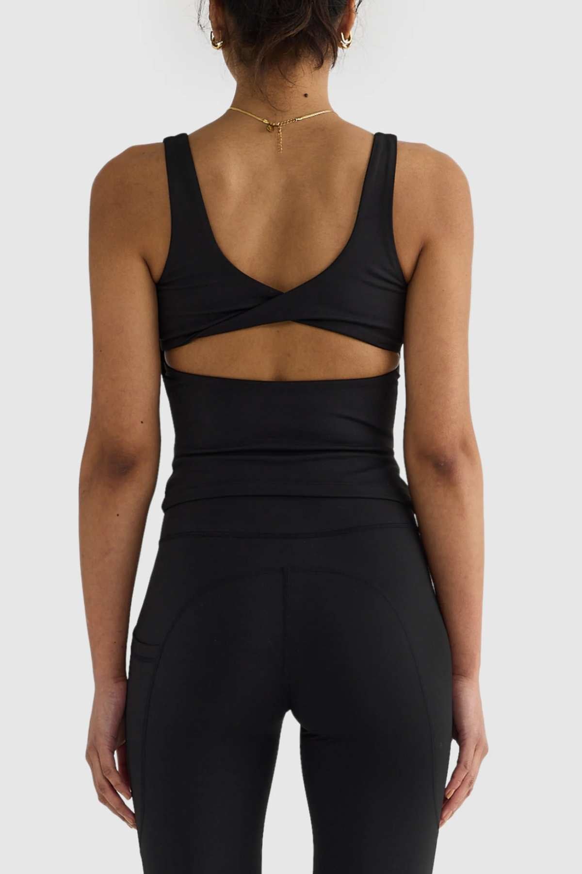 A woman confidently displays the ORTC Eva Twist Back Reversible Top, a stylish black sleeveless piece with a crisscross back, paired with matching leggings. Her look is elevated by a gold necklace and hoop earrings. Standing against a plain background, she highlights both her sense of style and the top's versatile dual-wear design.