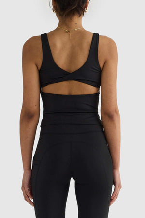 A woman confidently displays the ORTC Eva Twist Back Reversible Top, a stylish black sleeveless piece with a crisscross back, paired with matching leggings. Her look is elevated by a gold necklace and hoop earrings. Standing against a plain background, she highlights both her sense of style and the top's versatile dual-wear design.