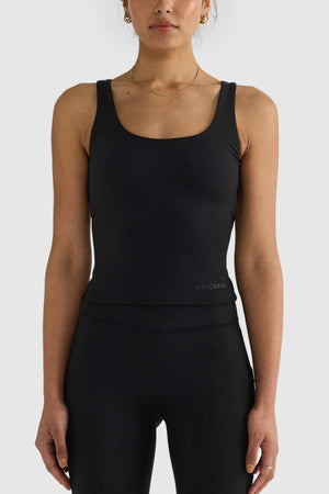 A person stands against a plain background wearing the ORTC Eva Twist Back Reversible Top in black along with matching black leggings. They accessorize with gold hoop earrings and a delicate necklace. The ensemble is form-fitting and sporty, ideal for versatile styling.