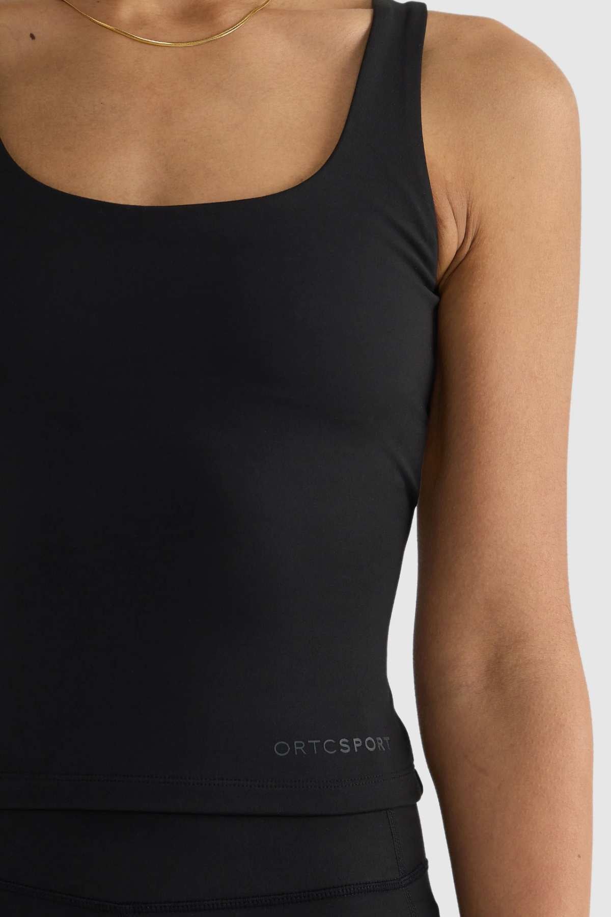 A person is wearing the ORTC Eva Twist Back Reversible Top, a black sports tank featuring "ORTCSPORT" near the hem. This top offers a dual-wear design that accentuates the upper torso against a neutral background.