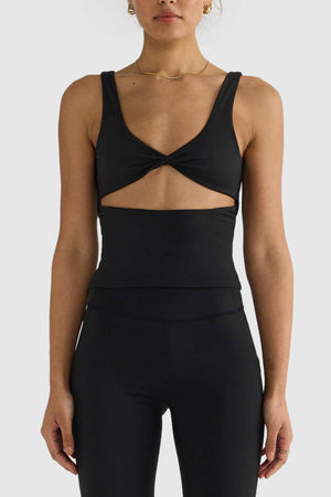 A person wearing the ORTC Eva Twist Back Reversible Top, a sleek black sleeveless piece with a twisted front design, paired with high-waisted leggings stands confidently against a plain gray background. This versatile top offers dual-wear design possibilities while maintaining an effortlessly chic look.