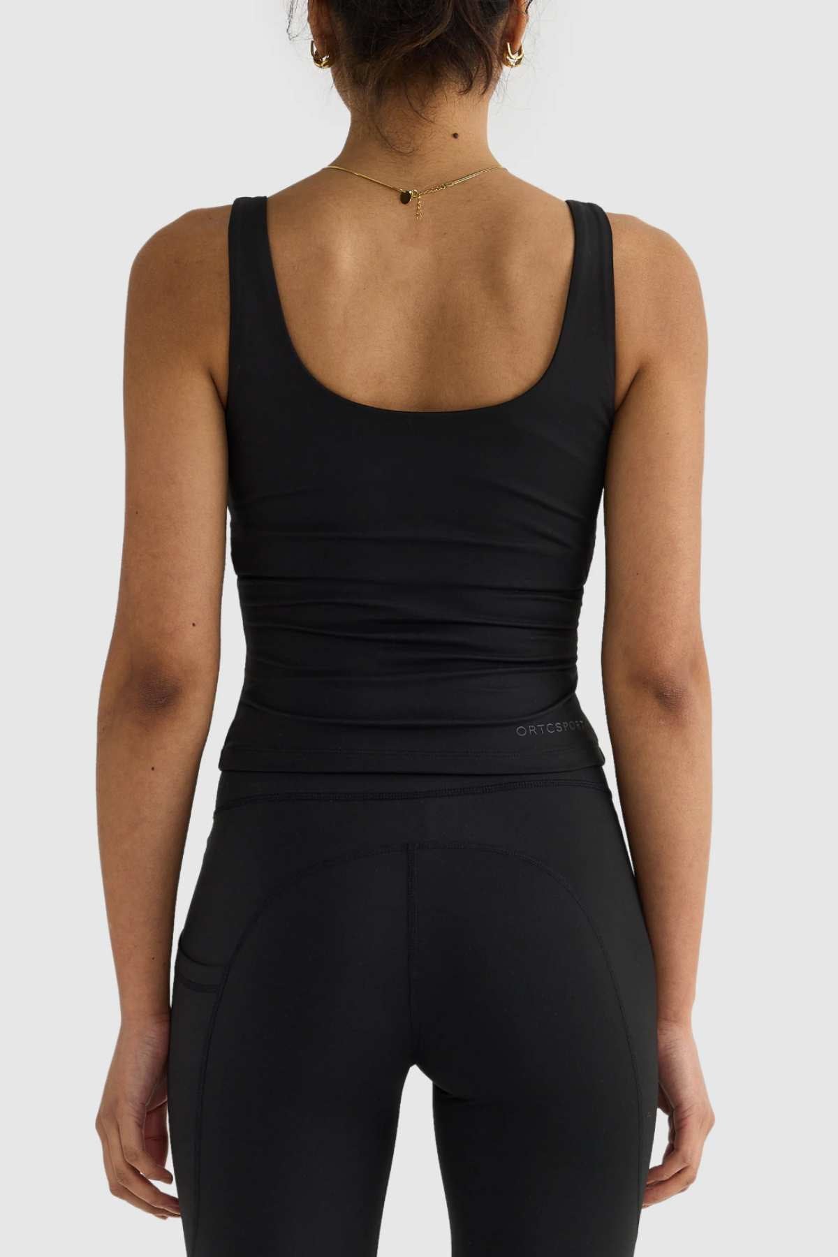 A woman is standing with her back to the camera in a sleek black ORTC Eva Twist Back Reversible Top and leggings featuring a dual-wear design. A delicate necklace graces her neckline, while her hair is elegantly tied up. The plain light grey background complements her sophisticated style.