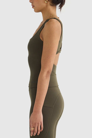 An individual dressed in an olive green activewear outfit is pictured in profile against a light background. They are wearing the ORTC Eva Twist Back Reversible Top, crafted from ultra-soft double-brushed fabric with a crossover back design, along with matching leggings. Their accessories consist of earrings and a ring.