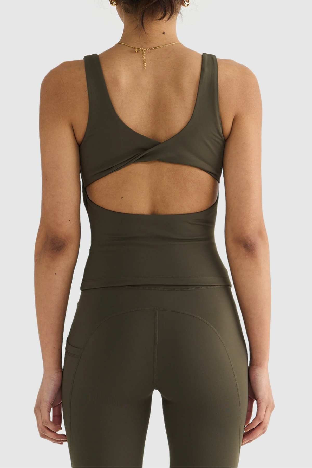A person is viewed from behind wearing the ORTC Eva Twist Back Reversible Top in olive green, accompanied by matching high-waisted leggings. The ultra-soft, double-brushed fabric of the top showcases a twist detail and cutout. With short curly hair and a necklace, their style exudes modern elegance.