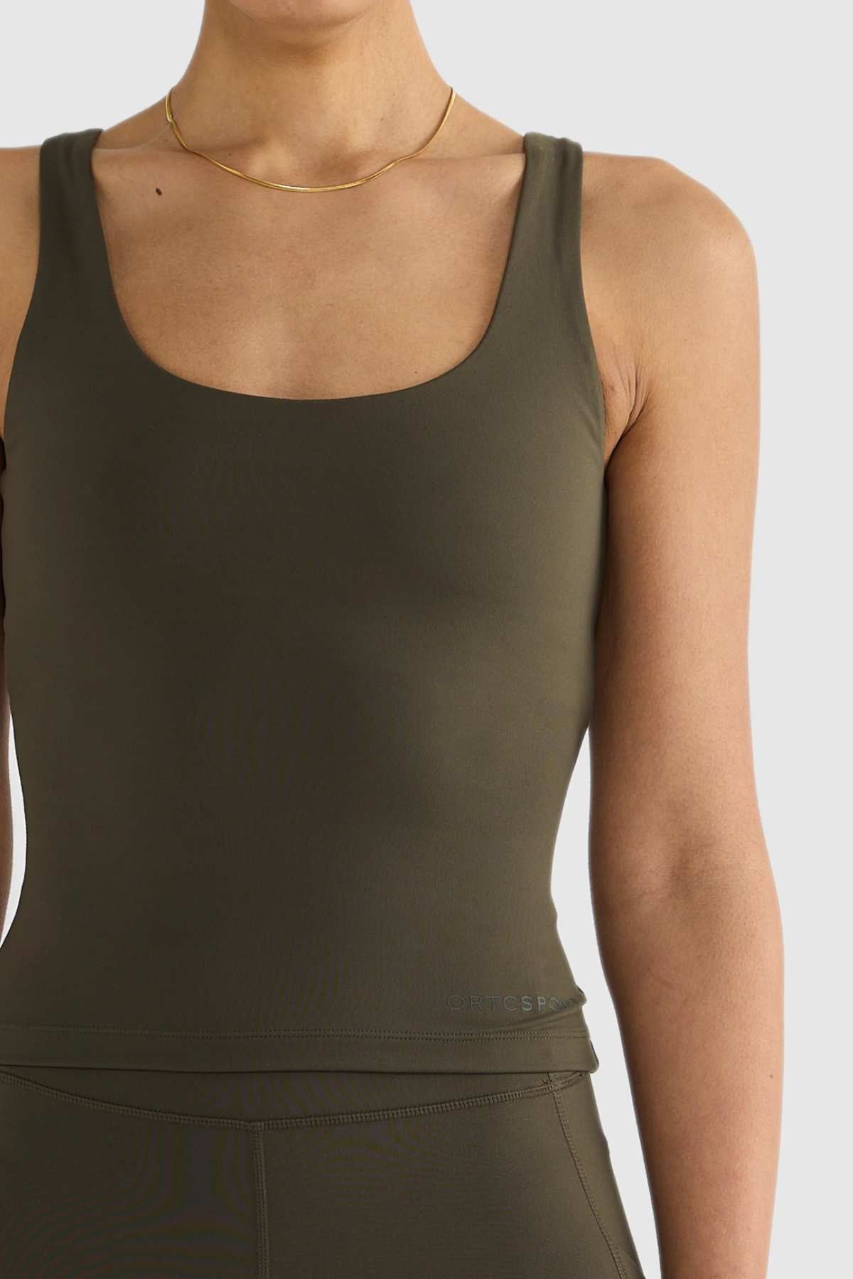 A person is depicted in an olive-green sleeveless bodysuit made from ultra-soft double-brushed material, paired with a delicate gold necklace, set against a simple backdrop. The ensemble mirrors the form-fitting and sleek design of the ORTC Eva Twist Back Reversible Top, emphasizing a minimalist and sporty aesthetic.