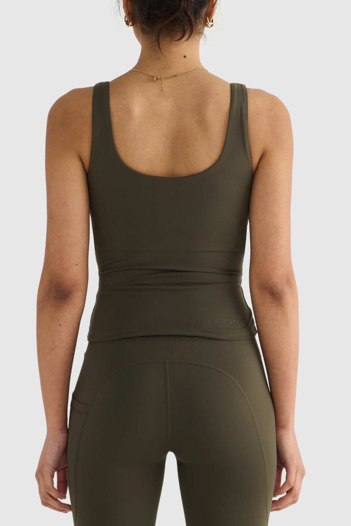 A person with dark hair in a bun is wearing an olive green ORTC Eva Twist Back Reversible Top and leggings, crafted from ultra-soft double-brushed fabric. The photo, captured from behind against a light gray background, beautifully showcases the outfit's dual-wear design.