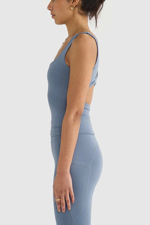 A person in a blue ORTC Eva Twist Back Reversible Top and leggings, crafted from double-brushed fabric, stands in profile against a white background. They wear gold hoop earrings, with dark hair elegantly tied up.