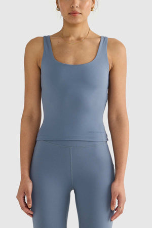 A person wearing a blue ORTC Eva Twist Back Reversible Top paired with matching leggings stands against a light background. With an elegant posture and arms at their sides, they accessorize with a simple necklace and earrings, highlighting the exquisite style of this reversible piece made from double-brushed fabric.