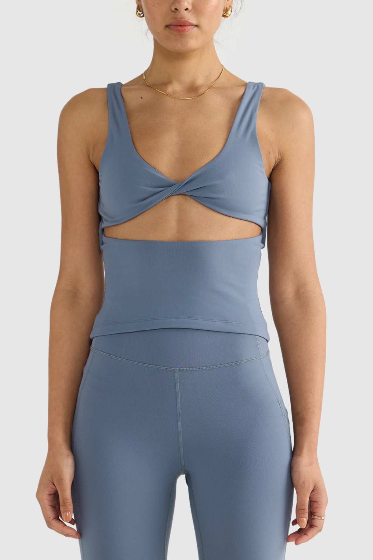 A person wearing a matching blue sports top and leggings stands against a plain, light background. The ORTC Eva Twist Back Reversible Top showcases a twist detail at the bust and a small cutout below, crafted from soft double-brushed fabric for ultimate comfort.