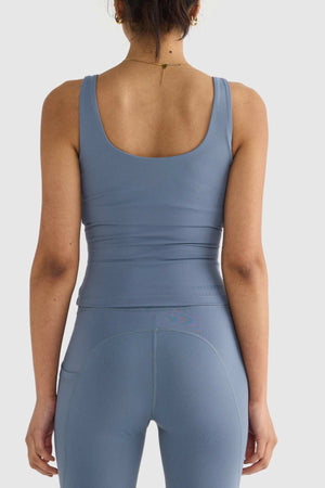 A person wearing a coordinated blue athletic tank top and leggings, featuring the ORTC Eva Twist Back Reversible Top design, stands with their back facing the camera. They have a thin gold chain necklace and their hair tied up, against a plain white background.