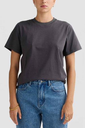 Dressed in a plain ORTC Everyday T-Shirt crafted from soft, mid-weight cotton, the person pairs it with blue jeans. Against a light gray background, they stand with hands resting at their sides. The relaxed fit tee is tucked in and complemented by a bracelet and earrings for added style.