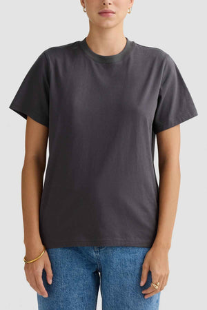 A person wearing a relaxed fit ORTC Everyday T-Shirt in plain dark gray pairs it with blue jeans while standing against a light gray background. Their hands rest casually by their sides, highlighting the gold hoop earrings and bracelets. Made from soft mid-weight cotton, this ensemble exudes an effortlessly casual vibe.