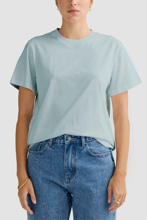 A person adorned in a stylish light green ORTC Everyday T-Shirt paired with blue jeans showcases both style and comfort. They complement their look with a gold bracelet and small hoop earrings, all set against a simple white background.