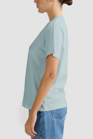 A person showcases effortless style wearing an ORTC Everyday T-Shirt combined with blue jeans, set against a plain background. Their short hair enhances the comfortable appearance, while small hoop earrings and rings add a touch of subtle flair.
