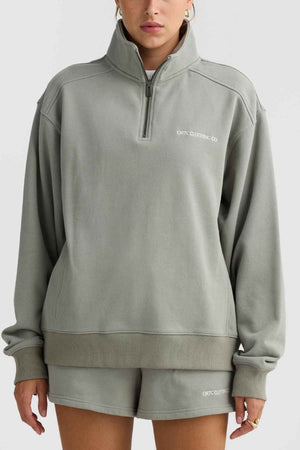 An individual dressed in the ORTC Lena Quarter Zip, a light gray-green terry cotton sweatshirt paired with matching shorts. The sweatshirt features a high collar adorned with the "ORTC CLOTHING CO." branding, which also appears on the shorts. They are set against a plain gray backdrop.