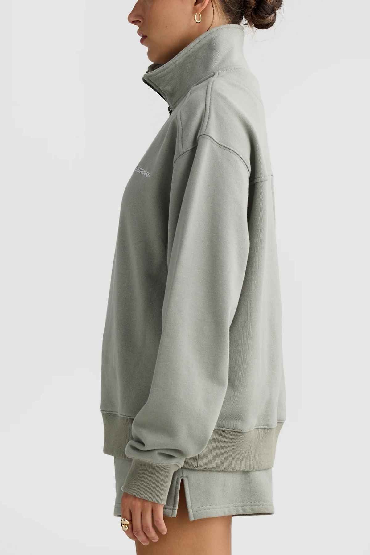 A person stands in profile against a plain background, wearing the ORTC Lena Quarter Zip sweatshirt and shorts in light green. The sweatshirt showcases a high collar and ribbed cuffs. The individual, with dark hair tied back, sports hoop earrings and a ring.