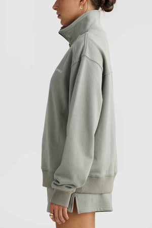 A person stands in profile against a plain background, wearing the ORTC Lena Quarter Zip sweatshirt and shorts in light green. The sweatshirt showcases a high collar and ribbed cuffs. The individual, with dark hair tied back, sports hoop earrings and a ring.