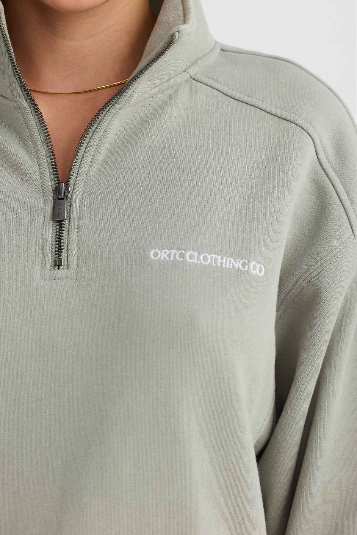 A person is wearing the ORTC Lena Quarter Zip, a terry cotton sweatshirt in light green featuring a small embroidered "ORTC CLOTHING CO" text on the left chest. They accessorize with a simple gold necklace against a plain white background.