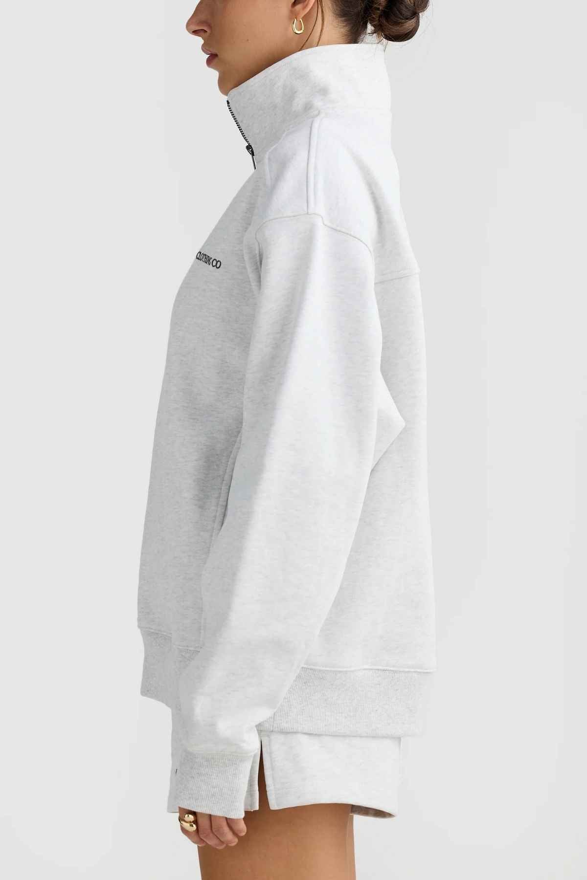 A person is shown in profile wearing the ORTC Lena Quarter Zip sweatshirt in light gray, which is designed with an ORTC branded metal zipper. Made from Terry Cotton, this piece offers long sleeves and a loose fit. The ensemble is complemented by small hoop earrings against a plain white background.