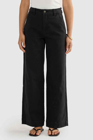 Wearing the relaxed-fit, high-waisted ORTC Lottie Relaxed Twill Pants with their wide-leg design, an individual effortlessly showcases style. Complemented by a basic white top and black sandals set against a light gray backdrop, the look is finished with a gold bangle adorning their left wrist.