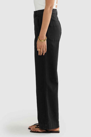 A person wearing a white shirt and ORTC Lottie Relaxed Twill Pants is standing in profile against a simple backdrop. They have bracelets on their wrist and are dressed in sandals, highlighting the relaxed fit style.