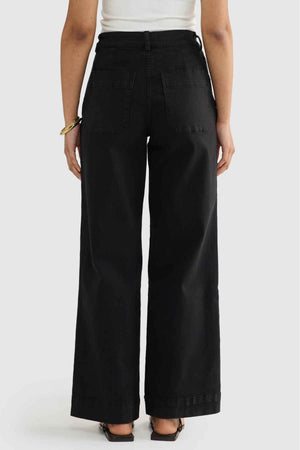 Someone wearing the ORTC Lottie Relaxed Twill Pants is standing with their back facing the camera, highlighting a high-waisted, wide-leg relaxed fit. They have paired it with a white top and black sandals, and are accessorizing with a bracelet on their right wrist.