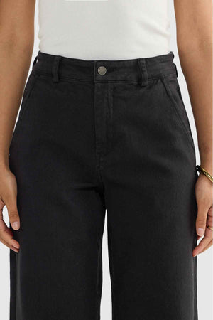 A person stands confidently, their arms relaxed at their sides, dressed in high-waisted ORTC Lottie Relaxed Twill Pants in black paired with a crisp white sleeveless top. The spotlight is on the relaxed fit and stretch cotton twill fabric, showcasing the meticulous detailing around the waist and pockets.