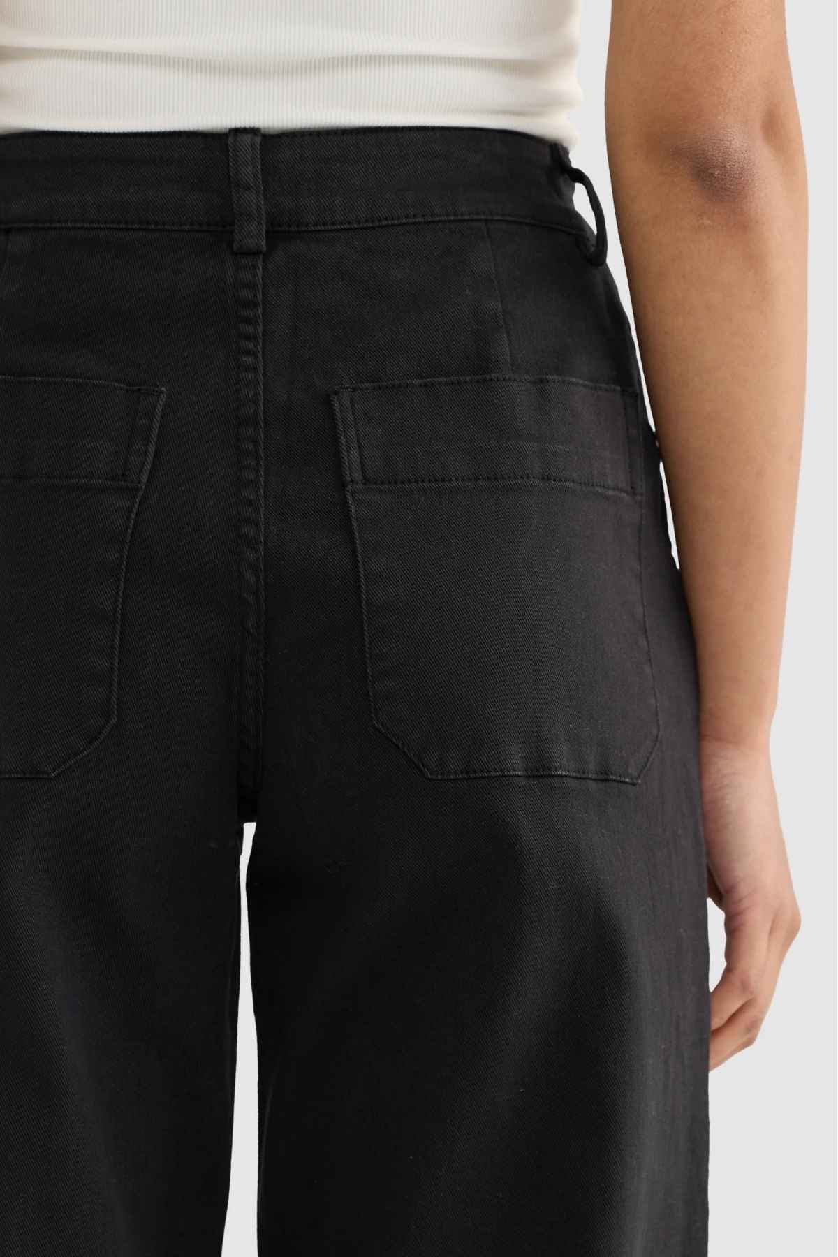 A person is wearing black ORTC Lottie Relaxed Twill Pants with large back pockets and a relaxed fit, paired with a white sleeveless top that is visible from the back. The smooth stretch cotton twill enhances comfort against the plain light gray background.