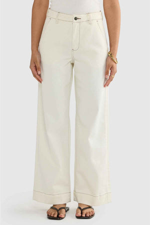 A person is wearing the Australian-designed ORTC Lottie Relaxed Twill Pants in a high-waisted, wide-leg cream style made from stretch cotton twill, paired with a white top. Their hands are resting by their sides, and they have completed the look with black sandals and a gold bracelet.