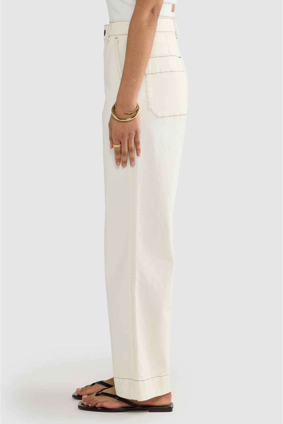 A person in ORTC Lottie Relaxed Twill Pants, designed in Australia, stands sideways wearing a white top. Their hands rest calmly at their sides, decorated with bracelets. They are wearing black flip-flops against a light gray background.