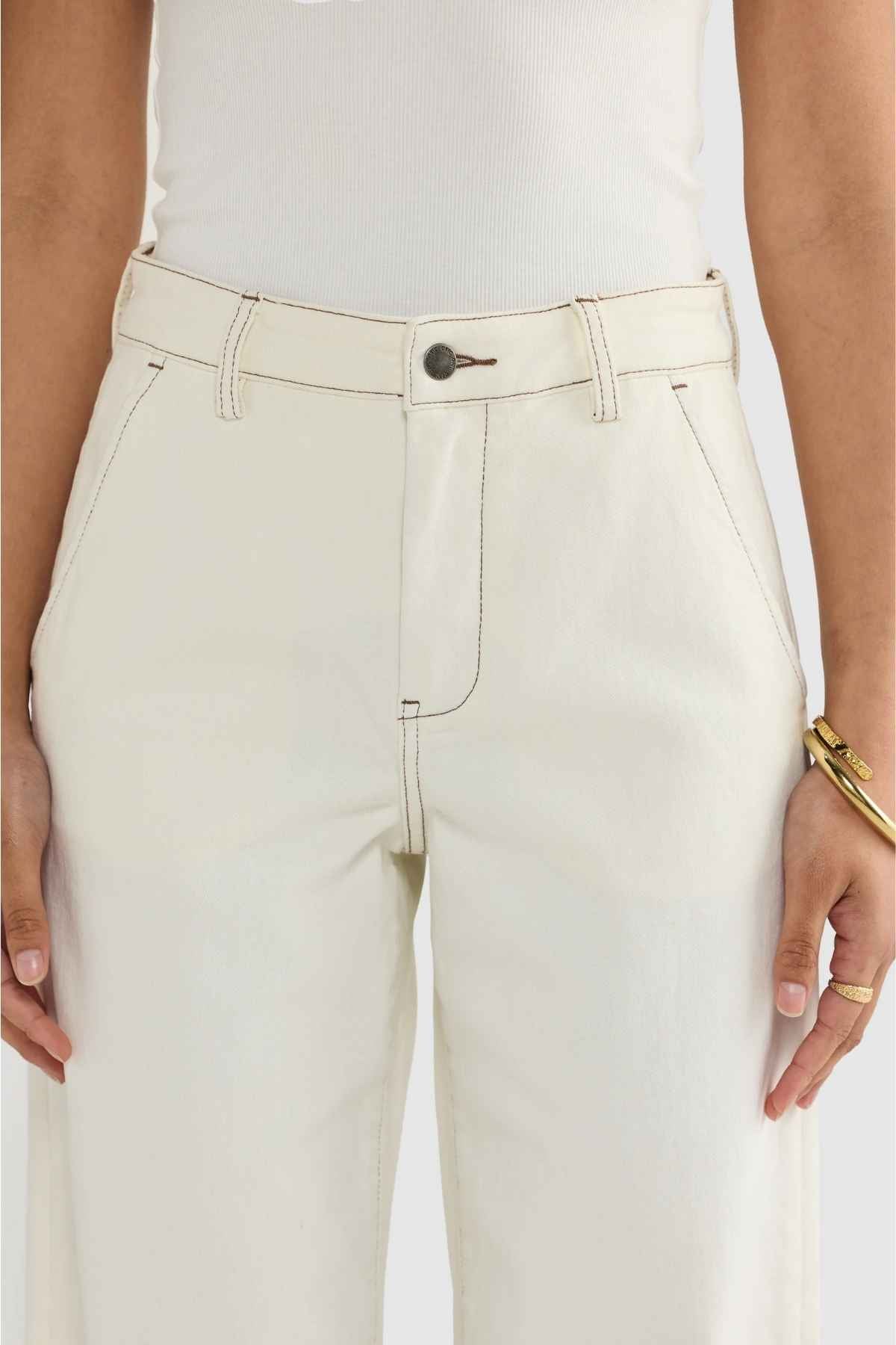A person wearing the ORTC Lottie Relaxed Twill Pants in cream, crafted from Australian-designed stretch cotton twill. These pants feature front pockets and a button closure, paired with a white top tucked in. Accessories include a gold bracelet and ring, with the image focusing on the waist and upper pants area.