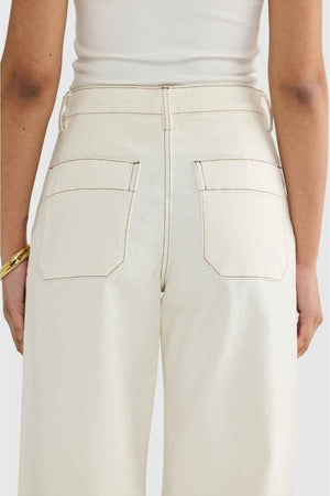 A person is shown from the back wearing ORTC Lottie Relaxed Twill Pants, crafted from Australian-designed stretch cotton twill, in a cream color. The high-waisted jeans feature two back pockets and are paired with a white sleeveless top. The look is complemented by a gold bracelet on their right wrist.