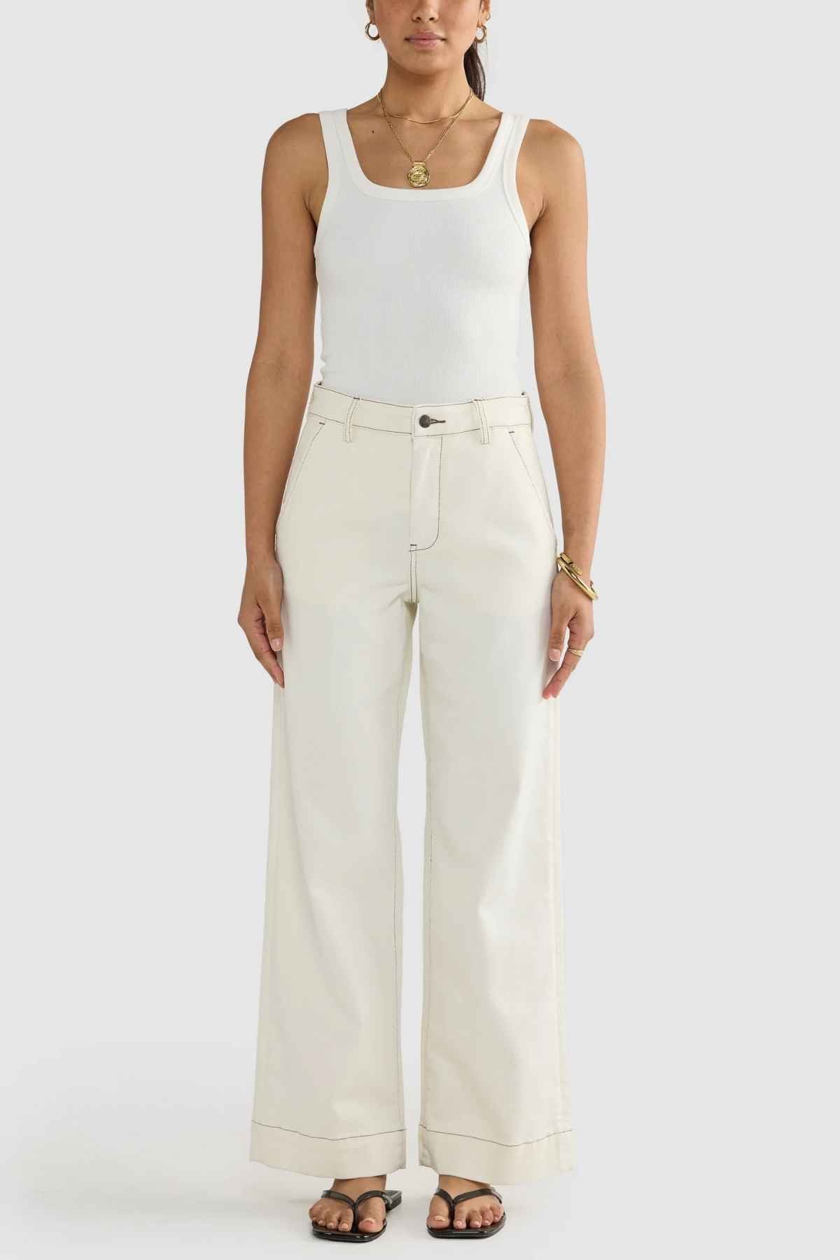 A person wearing the ORTC Lottie Relaxed Twill Pants in cream, paired with a white tank top, is standing against a plain background. They complete the look with black sandals and accessorize with bracelets and a necklace, their hands resting by their sides. This ensemble showcases the effortless elegance of Australian-designed stretch cotton twill.