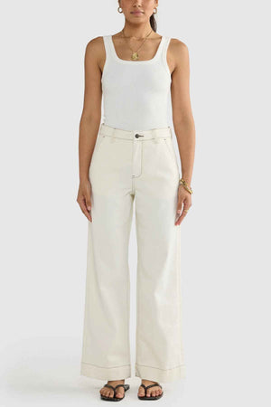 A person wearing the ORTC Lottie Relaxed Twill Pants in cream, paired with a white tank top, is standing against a plain background. They complete the look with black sandals and accessorize with bracelets and a necklace, their hands resting by their sides. This ensemble showcases the effortless elegance of Australian-designed stretch cotton twill.