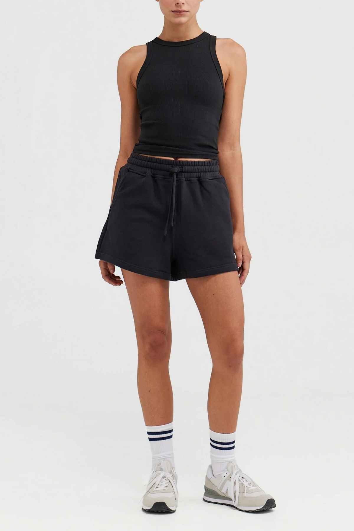 A person dressed in an ORTC Mimi Rib Tank and black shorts poses against a white background. They're sporting white sneakers paired with white socks featuring black stripes. The tank's ribbed knit fabric makes it ideal for layering, as they stand with their left hand at their side while their right hand rests on their hip.