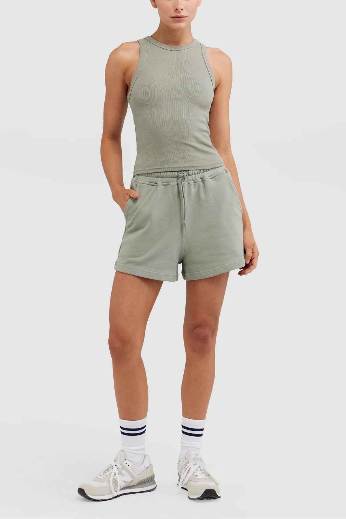 Dressed in a sleeveless ORTC Mimi Rib Tank paired with matching green shorts, an individual stands with one hand in a pocket. The ribbed knit fabric adds texture to the outfit. They complete the look with white socks featuring black stripes and beige sneakers, posing against a plain white background.