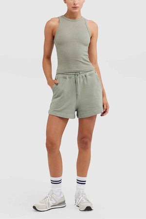 Dressed in a sleeveless ORTC Mimi Rib Tank paired with matching green shorts, an individual stands with one hand in a pocket. The ribbed knit fabric adds texture to the outfit. They complete the look with white socks featuring black stripes and beige sneakers, posing against a plain white background.