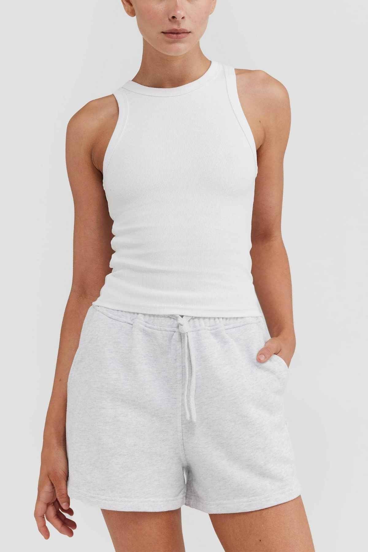 A person poses against a simple backdrop, showcasing the ORTC Mimi Rib Tank—an ideal layering piece made from ribbed knit material—complemented by light gray drawstring shorts. They have their left hand in one pocket while their right arm rests at their side.