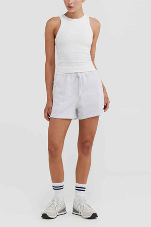 A person is pictured against a plain background, dressed in an ORTC Mimi Rib Tank in white, with light gray shorts and sneakers. The ensemble is finished off with white socks adorned with blue stripes, making it a perfect layering choice for any casual occasion.