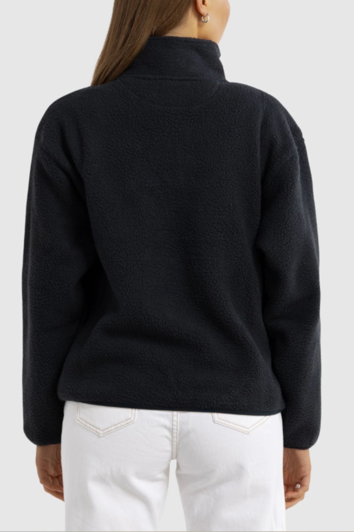 A woman with brown hair is seen from behind wearing the ORTC Sherpa Fleece Quarter Zip in black, featuring a high collar, paired with white jeans against a plain light gray background.