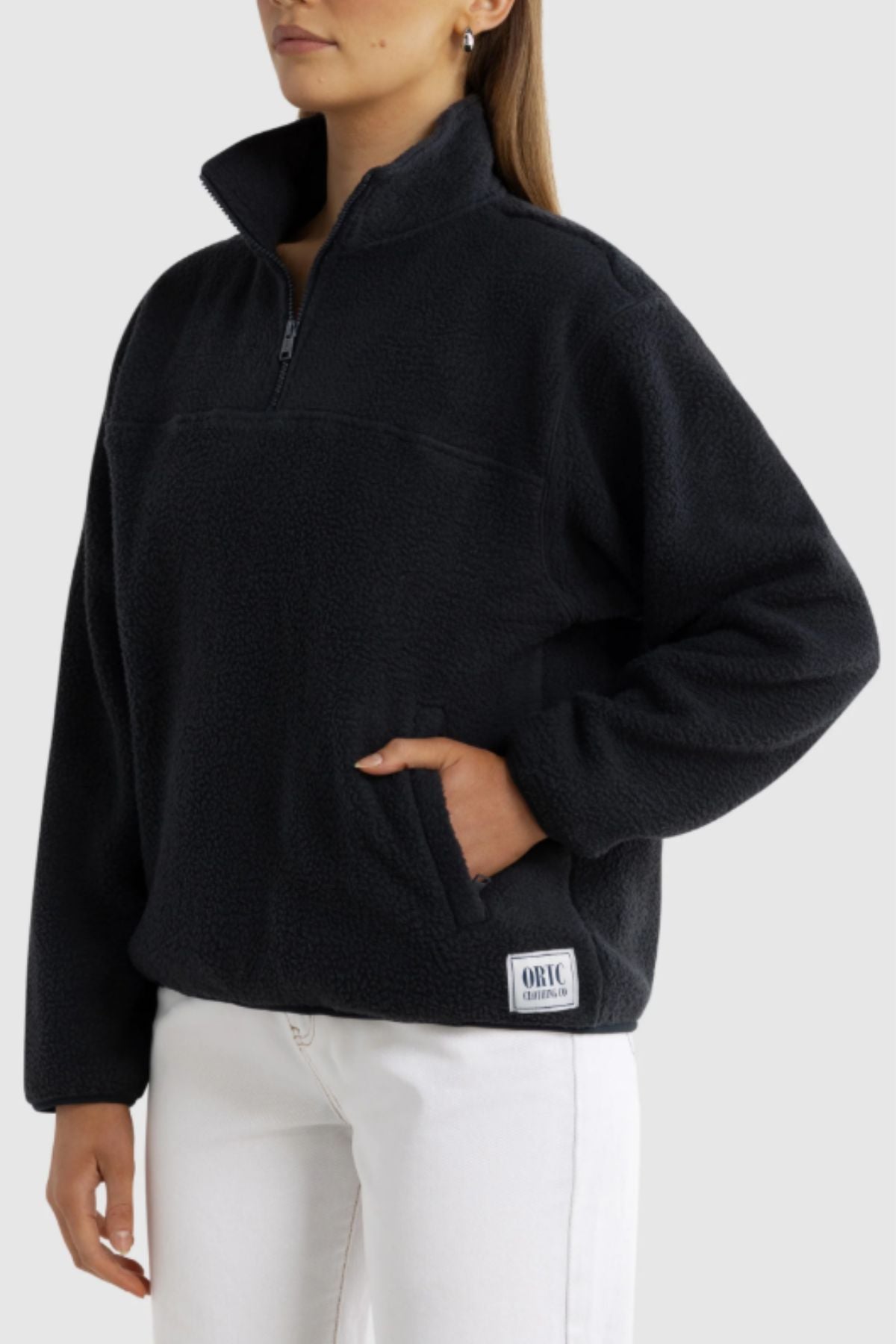 Someone wearing the ORTC Sherpa Fleece Quarter Zip in black, featuring a front pocket and a small rectangular patch near the hem, paired with white pants against a plain light gray background.
